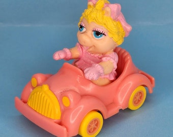Pode incluir: Um carro de brinquedo rosa com uma grelha amarela e rodas amarelas. Um personagem de porco com cabelo loiro está sentado no banco do motorista.