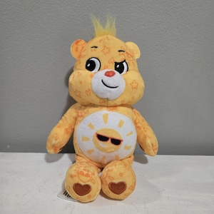 May include: A yellow Care Bear soft toy with a sun design on its tummy. The bear has a playful expression, with a wink and a tuft of yellow hair. The bear's body is covered in star and heart patterns. The sun emblem has sunglasses.