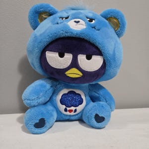 May include: A blue plush bear in a dark purple penguin costume. The bear has a sad expression and a glittery cloud and rain design on its belly. The penguin costume features a yellow beak and white eyes.