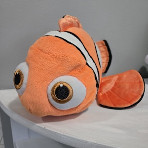 May include: Orange plush clownfish with white stripes, large white eyes with gold and black details, and a large orange fin. The plush toy is a soft, cuddly representation of a popular animated character.