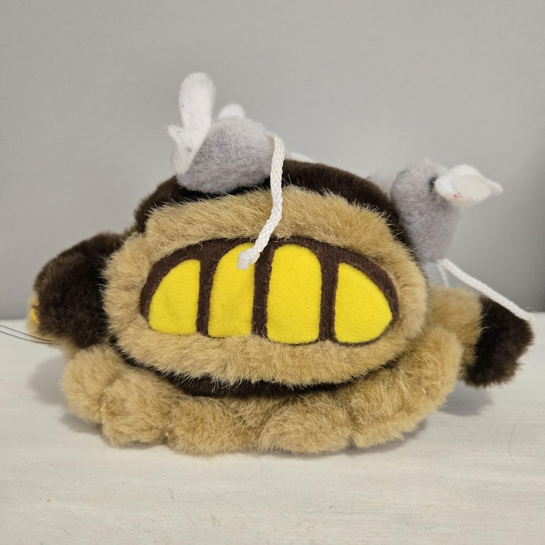 May include: A plush toy resembling a cartoon character, featuring a tan body with brown accents and large yellow teeth. Two gray mouse-like figures with white ears are attached to the top with white string.