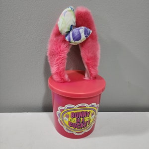 May include: Pink plastic bucket with a pink fuzzy bunny ear headband with a floral bow on top. The bucket has the words "Bunny in a Bucket" printed on it.