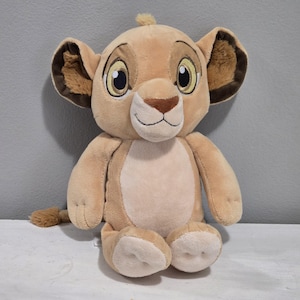 May include: A soft toy lion cub in shades of tan and beige. The plush animal has large eyes, a small nose, and a smiling mouth. The ears are lined with darker brown fabric. The toy is sitting upright with its tail visible.