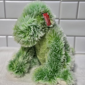Op de afbeelding: Groen pluche knuffeldier dat zit met een rood label met de tekst "Aurora" erop.