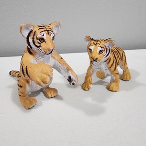 May include: Two plastic tiger figurines, one larger than the other. The larger tiger is orange and black with white stripes and a white chest. The smaller tiger is orange and black with white stripes and a white chest. Both tigers have black noses and black eyes.