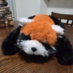 May include: A soft red panda toy with a furry texture. The toy is a mix of orange, black, and white, with a detailed face and realistic design. The toy is lying on a wooden surface.