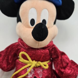 Pu&ograve; includere: Un Mickey Mouse di peluche che indossa una veste rossa e dorata con un cappello blu. Il giocattolo di peluche ha un naso nero e grandi occhi bianchi.