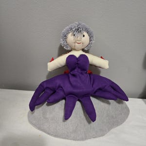 May include: A handmade doll with a gray pom-pom hairstyle, a cream-colored face, and a purple dress with tentacle-like extensions. The doll has red accents and is set on a gray, rounded base.