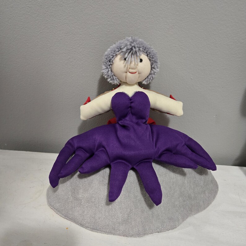 May include: A handmade doll with a gray pom-pom hairstyle, a cream-colored face, and a purple dress with tentacle-like extensions. The doll has red accents and is set on a gray, rounded base.