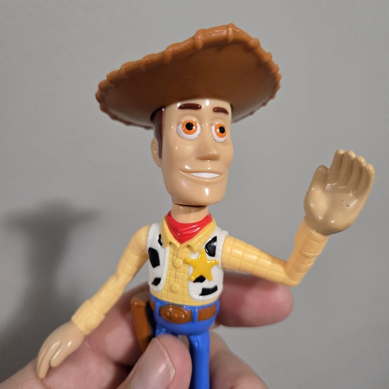 Pu&ograve; includere: Una figurina di plastica di Woody di Toy Story. Indossa un cappello da cowboy marrone, un gilet giallo con una stella da sceriffo e jeans blu. Sta salutando con la mano destra.