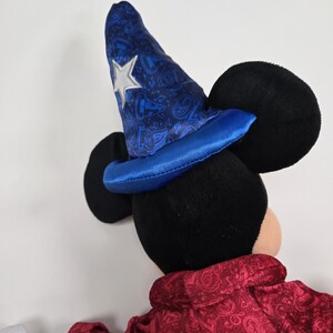 Pu&ograve; includere: Un Mickey Mouse di peluche che indossa un cappello da mago blu e argento con una tunica di velluto rosso. Il cappello ha una stella sul davanti e un motivo paisley.