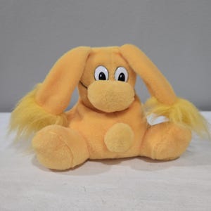 Può includere: Un peluche a forma di animale dei cartoni animati, principalmente arancione con dettagli gialli. Il giocattolo ha grandi orecchie flosce, un muso arrotondato e occhi bianchi con pupille nere. Il giocattolo è appoggiato su una superficie bianca.