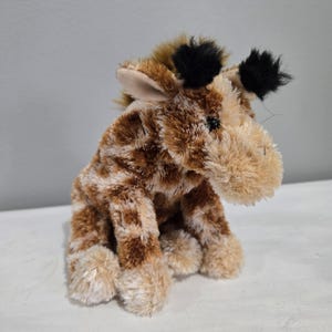 Op de afbeelding: Een pluche giraf met een bruin en beige gevlekt patroon. Het knuffeldier heeft een lichtbruin gezicht en poten, met zwarte plukjes op zijn hoofd. De giraf zit rechtop, met zijn hoofd iets opzij gedraaid.