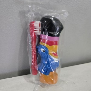 May include: A red toothbrush with white bristles and a blue and yellow character toothbrush holder, packaged in clear plastic. The toothbrush holder has a black top and a blue bird design.