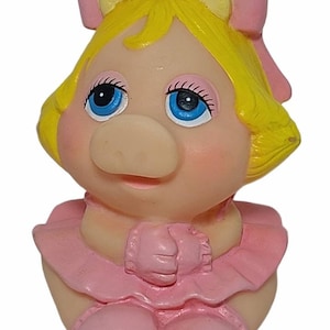 May include: A cartoon pig figurine with yellow hair, blue eyes, and a pink bow. The pig is wearing a pink dress. This figurine is likely a collectible or toy.