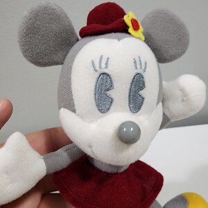 May include: A gray and white plush toy mouse wearing a red hat with a yellow flower. The mouse has embroidered eyes and a gray nose.