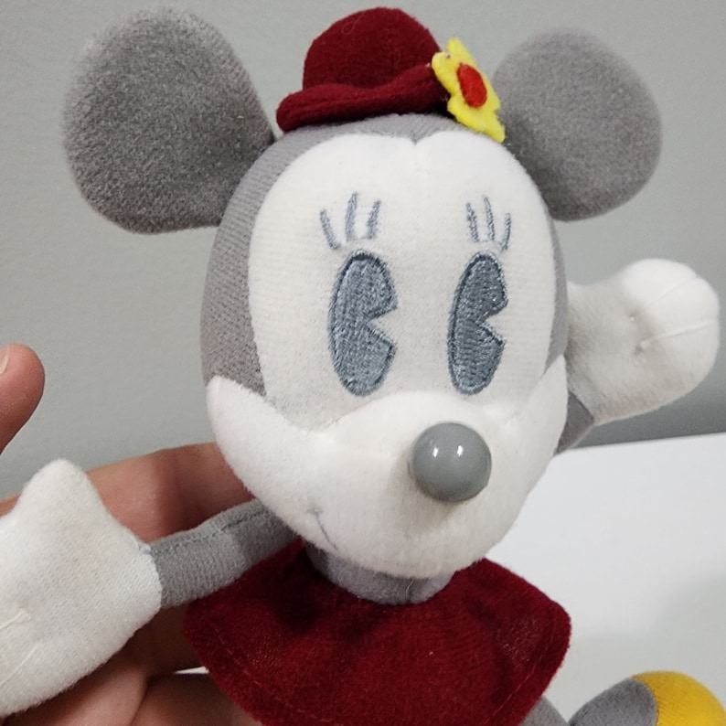 May include: A gray and white plush toy mouse wearing a red hat with a yellow flower. The mouse has embroidered eyes and a gray nose.