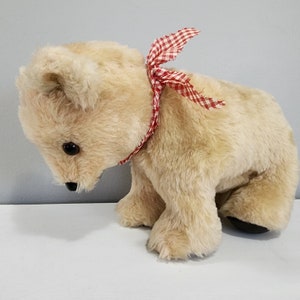Peut inclure: Un ours en peluche beige avec un nœud rouge et blanc à carreaux autour du cou. L'ours regarde vers le bas et a des yeux noirs.