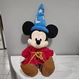 May include: A plush Mickey Mouse wearing a blue wizard hat with stars and a crescent moon. He is wearing a red robe with a gold rope tied around his neck.