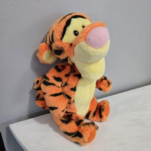 Könnte beinhalten: Ein Plüschtiger mit orangefarbenem Fell und schwarzen Streifen. Der Tiger hat ein gelbes Gesicht, Ohren und Bauch sowie eine rosa Nase. Das Spielzeug sitzt aufrecht, mit ausgestreckten Armen und Beinen.