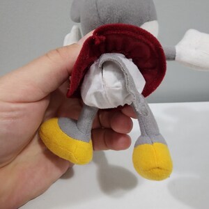May include: A gray plush toy with a red skirt and yellow feet. The toy is being held by a hand.