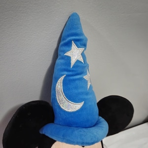 May include: A plush Mickey Mouse toy wearing a blue sorcerer hat with a silver crescent moon and stars.