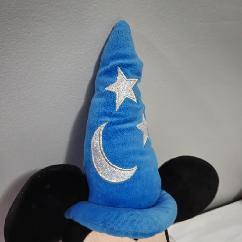May include: A plush Mickey Mouse toy wearing a blue sorcerer hat with a silver crescent moon and stars.