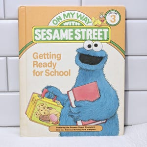Könnte beinhalten: Ein Vintage-Sesamstraße-Buch mit dem Titel "Getting Ready for School". Das Cover zeigt Krümelmonster, das ein rotes Buch und eine Lunchbox mit Keksen hält. Das Buch hat einen orangefarbenen und grünen Rand mit dem Text "On My Way With Sesame Street."