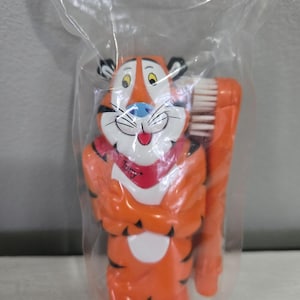 Könnte beinhalten: Orange und weiße Zahnbürste mit der Zeichentrickfigur Tony der Tiger. Die Zahnbürste befindet sich in einer durchsichtigen Plastiktüte. Die Figur hat einen roten Schal mit dem Namen "Tony". Die Zahnbürstenborsten sind weiß.