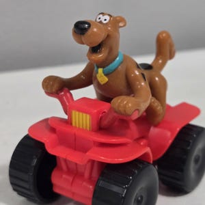 May include: A brown Scooby-Doo figurine with a light blue collar and yellow tag, riding a bright red four-wheeled all-terrain vehicle. The vehicle features a yellow front grille and four thick black tires with a textured tread pattern.