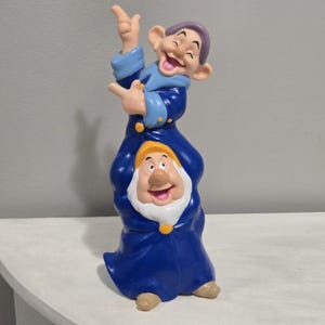 以下が含まれることがあります： 2人のキャラクターが重なった縦長のフィギュア。上のキャラクターは紫色の帽子、水色の袖、濃い青色のローブを着用。下のキャラクターは黄色の帽子、白いひげ、濃い青色のローブを着用している。全体的に滑らかな質感で、明るい表情をしている。