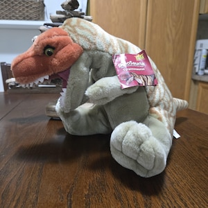 May include: A plush Tyrannosaurus dinosaur toy with a rust-colored head and a tan body featuring light brown stripes. The toy has an open mouth with white teeth and a red tag attached to its side that reads DinoSauria Tyrannosaurus.