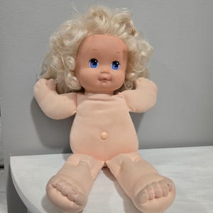 May include: A vintage baby doll with a soft, peach-coloured body and limbs. The doll has a plastic face with blue eyes and blonde, curly hair. The doll is sitting with its arms raised.