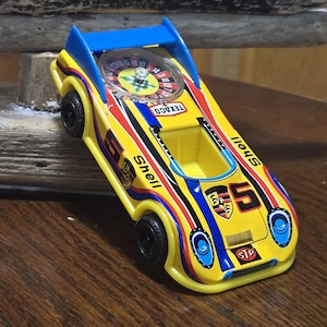 Könnte beinhalten: Ein gelbes Spielzeug-Rennauto mit blauen und roten Rennstreifen, das die Nummer 5, Porsche- und Shell-Logos sowie einen STP-Aufkleber trägt. Das Auto hat ein klares, kreisförmiges Rouletterad auf der Motorhaube und einen markanten blauen Heckflügel.