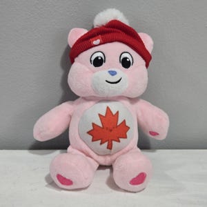May include: A pink plush bear wearing a red knitted hat with a white bobble. The bear has a white belly with a red maple leaf. The bear has black eyes, a blue nose, and a smiling mouth. The bear is sitting.