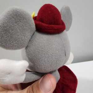 May include: A gray plush mouse wearing a red hat with a yellow star. The mouse has white wings and a red skirt.