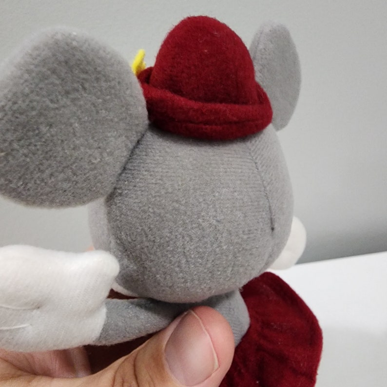 May include: A gray plush mouse wearing a red hat with a yellow star. The mouse has white wings and a red skirt.
