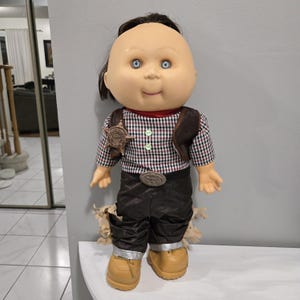 Könnte beinhalten: Eine Cabbage Patch Kid Puppe, die als Cowboy verkleidet ist. Die Puppe trägt ein kariertes Hemd, eine braune Weste, ein Sheriffstern-Abzeichen, eine braune Hose und braune Stiefel.