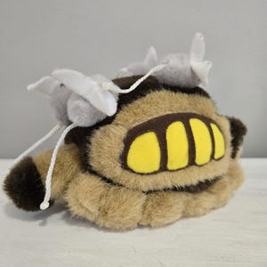 May include: A plush toy in the shape of a creature with a tan body, brown accents, and yellow details. The toy has two gray pom-poms with white accents and white string attached. The plush toy is on a white surface.