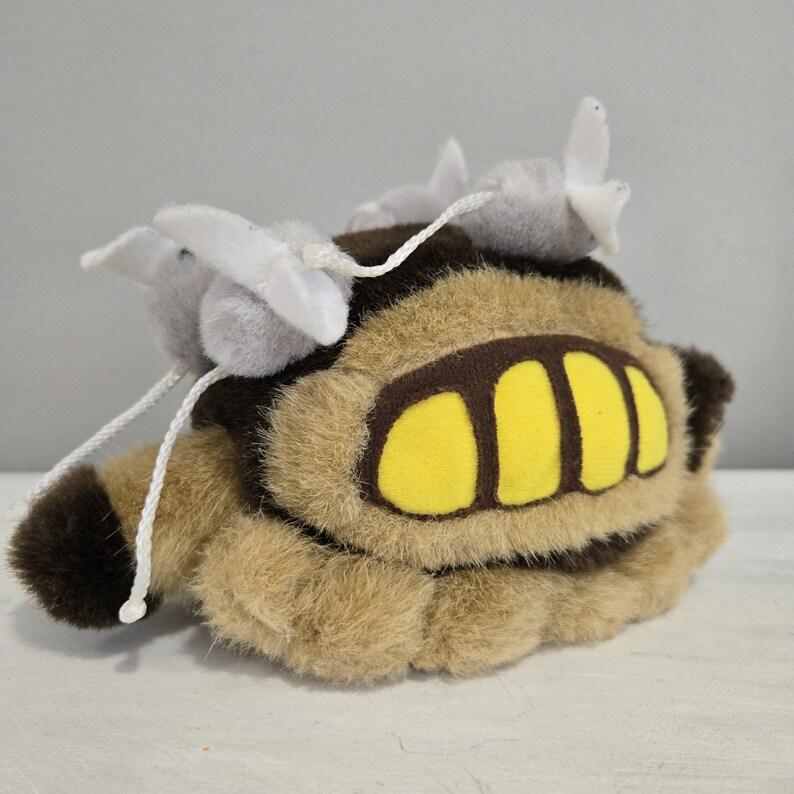 May include: A plush toy in the shape of a creature with a tan body, brown accents, and yellow details. The toy has two gray pom-poms with white accents and white string attached. The plush toy is on a white surface.