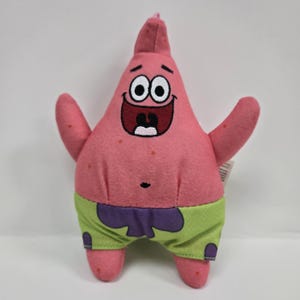 May include: Pink plush toy of Patrick Star from Spongebob Squarepants. Patrick is wearing green shorts with purple trim and has a big smile on his face.