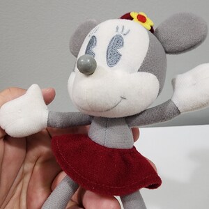 May include: A gray and white plush toy mouse wearing a red skirt and a yellow flower on its head. The mouse has embroidered eyes and a gray nose.