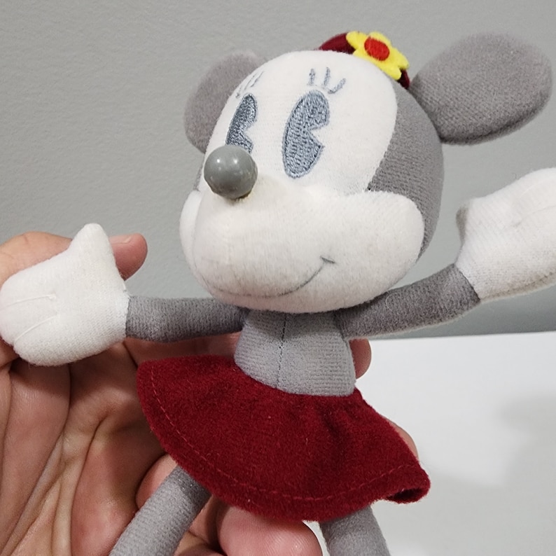May include: A gray and white plush toy mouse wearing a red skirt and a yellow flower on its head. The mouse has embroidered eyes and a gray nose.