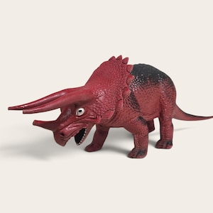May include: A plastic triceratops dinosaur figurine featuring a textured red body with black shading along the back and tail. The figure has three horns, a large frill, and an open mouth showing white teeth, standing on four legs.
