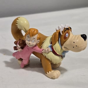 May include: A figurine depicting a child in pink pajamas and a teddy bear riding on the back of a Saint Bernard dog. The dog has a white and tan coat with a blue bow tie.