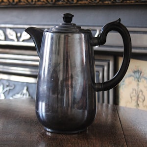 May include: A silver coffee pot with a black handle and a black knob on the lid. The pot is sitting on a wooden table.