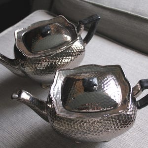 May include: Two silver teapots with hammered metal finishes and black handles. The teapots have a lid with a small knob on top.