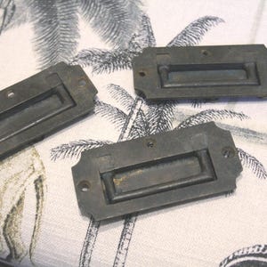 May include: Three antique brass drawer pulls with a rectangular shape and a recessed handle. The pulls have a worn, aged finish.