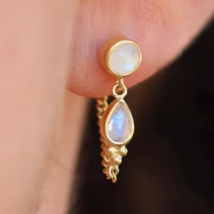 May include: Gold earrings with a teardrop-shaped moonstone and a round moonstone. The earrings are connected by a chain of small gold beads.