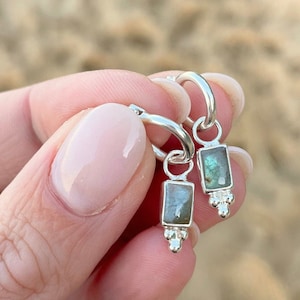 May include: A pair of silver hoop earrings with a dangling rectangular labradorite stone charm. Each charm has three small silver beads at the bottom.
